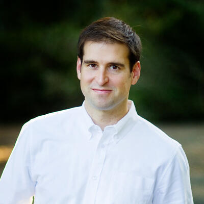 Man with brown hair in white button up shirt in front of greenery