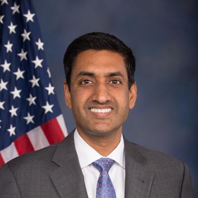 Dark haired man looks smiles directly at camera while in front of an American flag