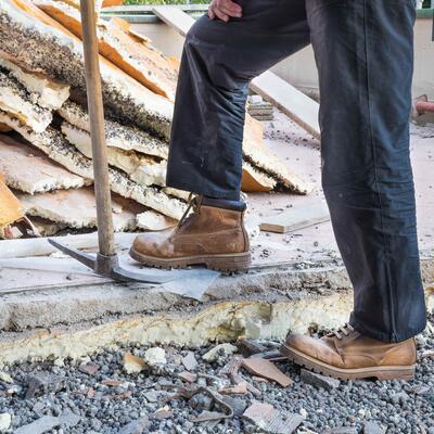 A photo of a the lower half of a worker who is wearing jeans and work boots and leaning against a pickaxe while doing construction work